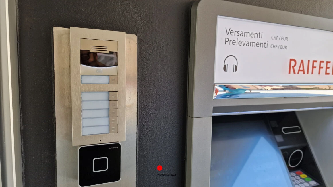 Videocitofono con telecamera e lettore di badge RFID installato a parete accanto a uno sportello bancomat Raiffeisen