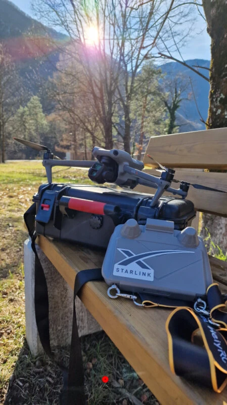 Kit Starlink e drone posizionati su una panchina in legno con sfondo montano e luce solare filtrata tra gli alberi