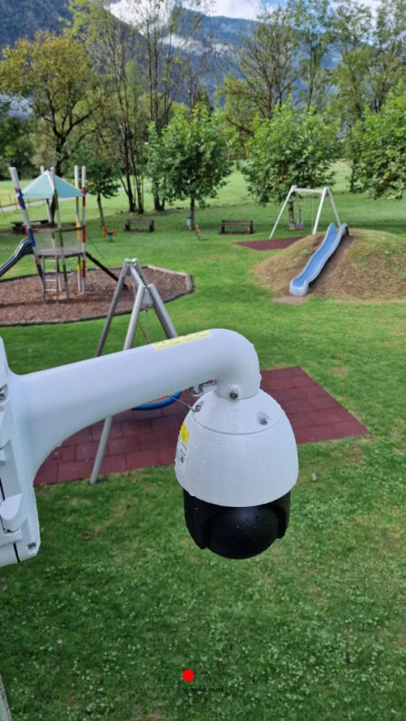 Telecamera di videosorveglianza PTZ montata vicino a un parco giochi con altalene e scivoli, immerso nel verde con sfondo montano
