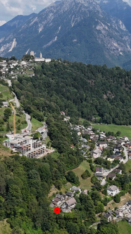 Vista aerea con drone di un centro abitato in area montana, con case immerse nel verde e panorama alpino sullo sfondo